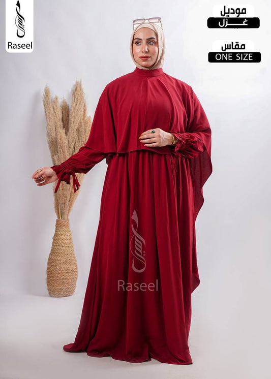 A woman modeling a deep red modest long-sleeve abaya dress with a high neck and a matching hijab, standing next to a decorative vase with dried grass. The image includes text overlays for 'Raseel' and 'ONE SIZE' in Arabic and English.