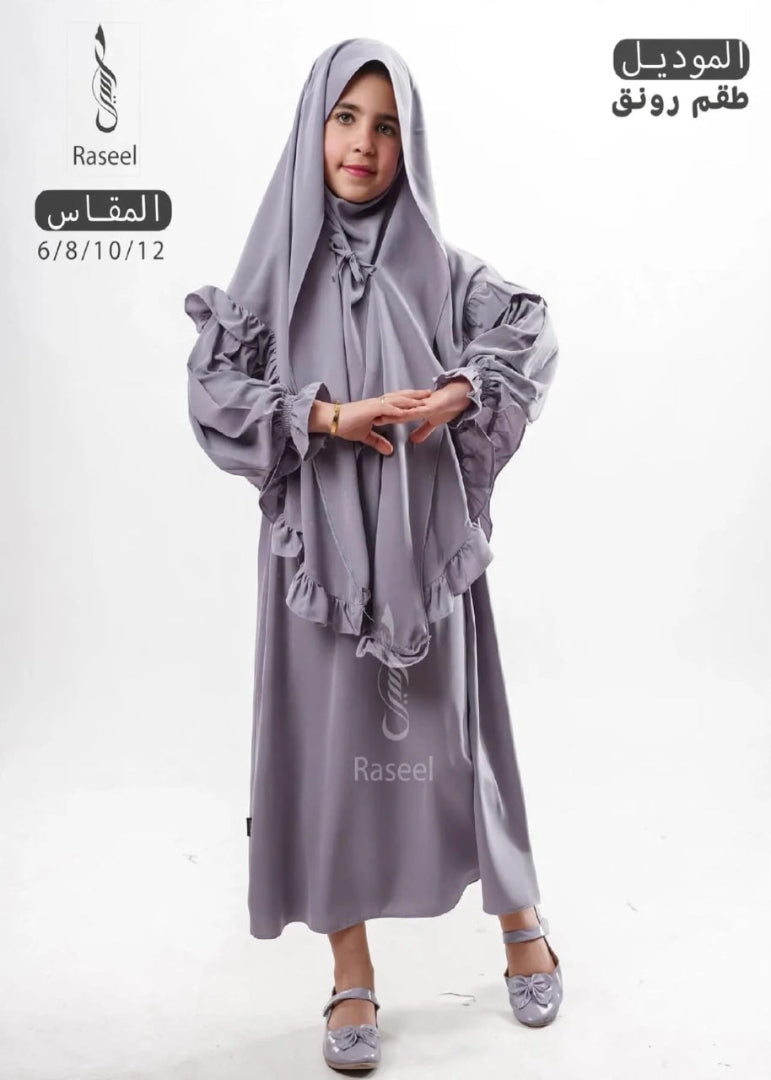 A young girl modeling a lavender-gray modest clothing set consisting of a long abaya dress with ruffled sleeves and a matching hijab, shown against a plain white background with Arabic text overlays.