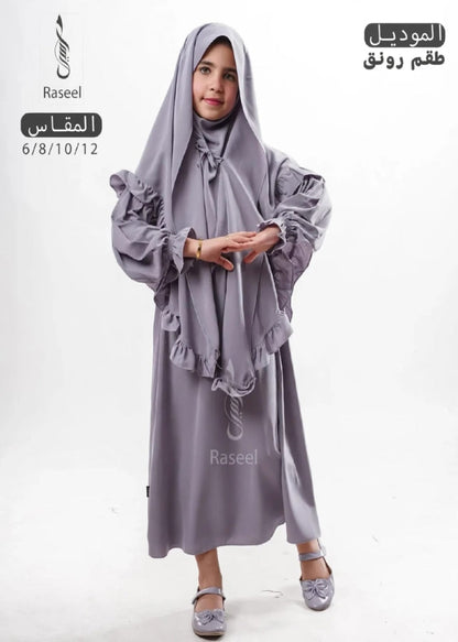 A young girl modeling a lavender-gray modest clothing set consisting of a long abaya dress with ruffled sleeves and a matching hijab, shown against a plain white background with Arabic text overlays.