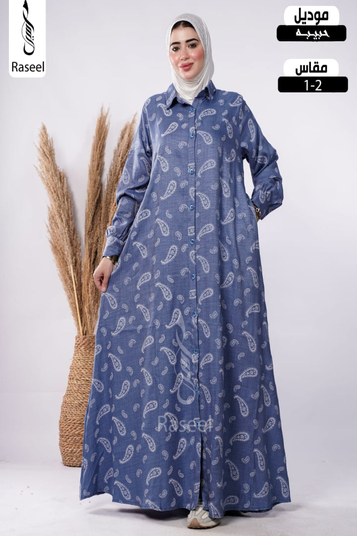 A woman wearing a long-sleeved blue button-front abaya with a white paisley pattern, paired with a light gray hijab, standing next to a decorative pampas grass basket. The image includes overlay text for 'Raseel' and Arabic text indicating 'Mouadil' and 'Mfas 1-2'.