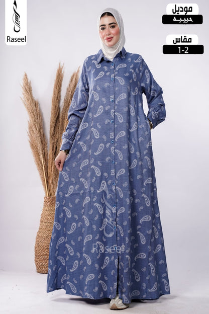 A woman wearing a long-sleeved blue button-front abaya with a white paisley pattern, paired with a light gray hijab, standing next to a decorative pampas grass basket. The image includes overlay text for 'Raseel' and Arabic text indicating 'Mouadil' and 'Mfas 1-2'.