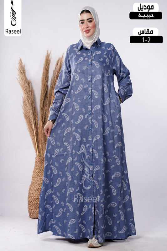 A woman wearing a long-sleeved blue button-front abaya with a white paisley pattern, paired with a light gray hijab, standing next to a decorative pampas grass basket. The image includes overlay text for 'Raseel' and Arabic text indicating 'Mouadil' and 'Mfas 1-2'.