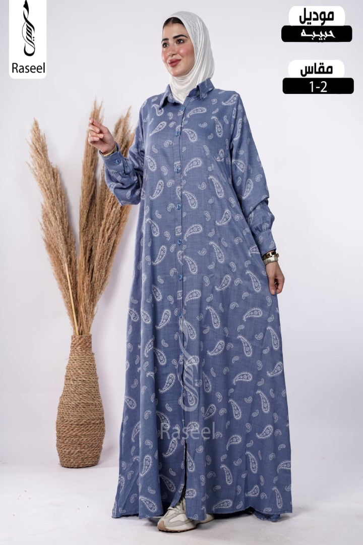 A woman wearing a long-sleeved, button-front abaya in a dusty blue paisley pattern, paired with a white hijab. She is standing next to a decorative vase of dried pampas grass against a white studio background.