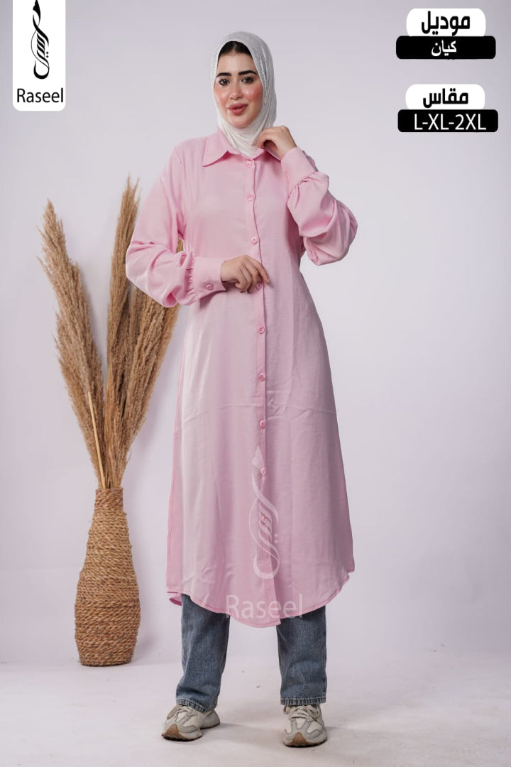 A woman modeling a light pink long-sleeve button-front abaya dress with a collar, paired with blue jeans and a light gray hijab, standing in front of a light gray studio background with a decorative pampas grass vase. Arabic text overlays indicate the product type and size range.