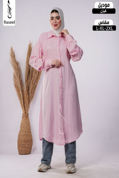 A woman modeling a light pink long-sleeve button-front abaya dress with a collar, paired with blue jeans and a light gray hijab, standing in front of a light gray studio background with a decorative pampas grass vase. Arabic text overlays indicate the product type and size range.