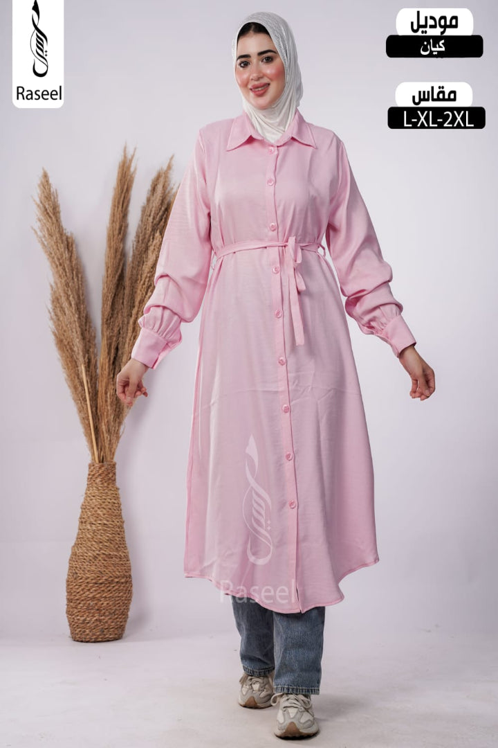 A woman wearing a light pink long-sleeve button-front tunic dress with a tie waist, styled with a white hijab, blue jeans, and sneakers. The image has 'Raseel' branding and Arabic text indicating the product is a tunic with a matching seat pad, sizes L-XL-2XL.