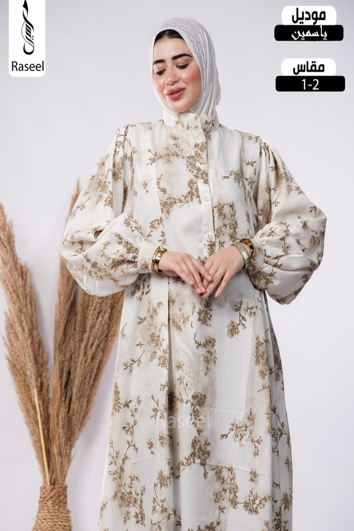 A woman wearing a long beige modest dress with a brown floral pattern and a matching hijab, standing against a light gray background with decorative dried grass on the left. Arabic text overlays on the right indicate the product type and size.